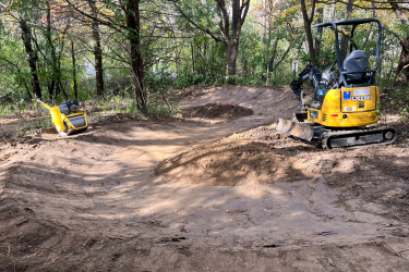 A construction site in a forested area featuring a yellow compact excavator and a yellow plate compactor on freshly leveled dirt. The environment is surrounded by trees with hints of autumn foliage. Carlson Park mountain bike trail.