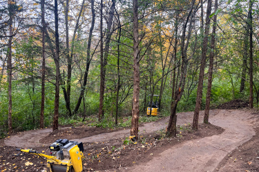 A wooded area with trees displays a newly constructed dirt trail that winds through the forest. In the foreground, a yellow compacting machine is resting on the ground, while another similar machine is visible further along the trail. The scene features a mix of greenery and fallen leaves, indicative of the autumn season. Carlson Park mountain bike trail.