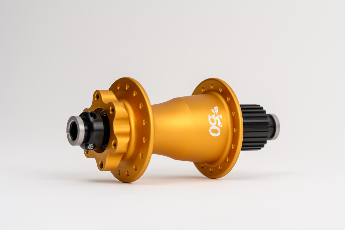 A close-up view of a Chris King Super Boost 6-bolt bicycle hub, featuring a shiny gold aluminum body with a black center. The hub has multiple drilled holes around the edge for spoke attachment and displays a white logo. The background is plain and light, highlighting the details and finish of the hub.