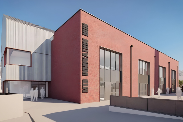 A modern brick building with distinctive architectural features, labeled "MIB Innovation Hub." The structure has large glass windows and a slanted roof design. In the foreground, several figures are walking, suggesting a busy atmosphere. The background features trees and additional buildings, under a clear blue sky.