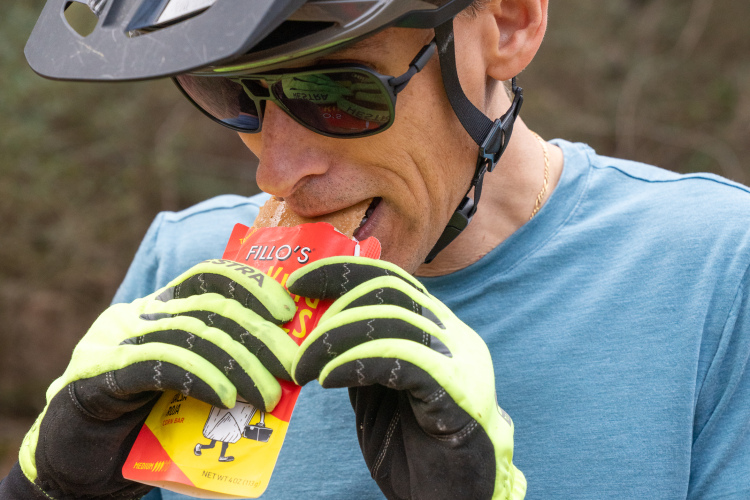 A cyclist wearing a black helmet and sunglasses is about to take a bite of a snack bar while standing outdoors. He is dressed in a blue shirt and has on bright yellow gloves. The background is nature, suggesting he is taking a break during a ride.