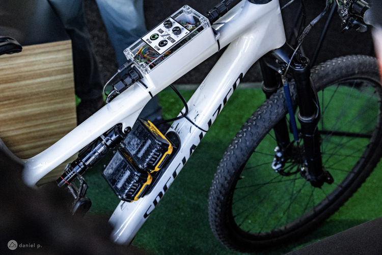 An electric mountain bike featuring additional electronic components mounted on the frame, including a control panel with buttons and lights. The bike frame is predominantly white, and two battery packs are attached to the lower section. The background shows a green grass-like surface, and parts of a person&rsquo;s legs can be seen next to the bike.