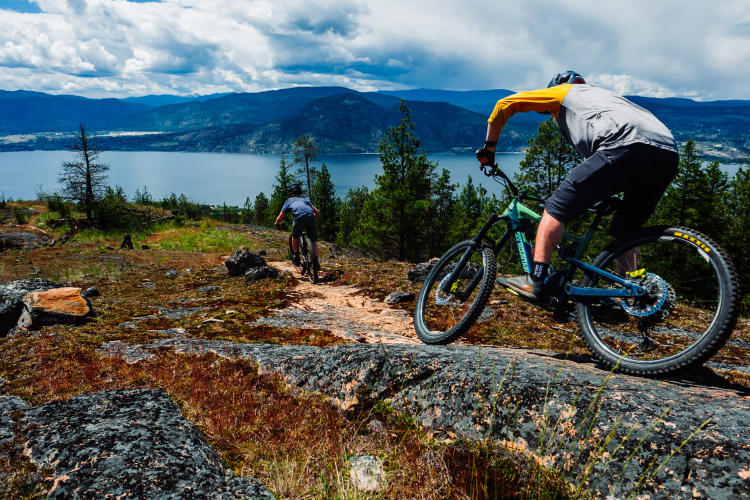 Two mountain bikers navigating a rocky trail with a scenic view of a lake and mountains in the background. The sky is partly cloudy, and the landscape features greenery and boulders.