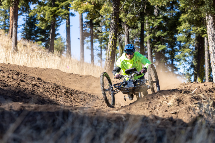 A cyclist in a bright yellow jacket riding a handcycle along a dirt trail, surrounded by tall trees and dust being kicked up from the ground.