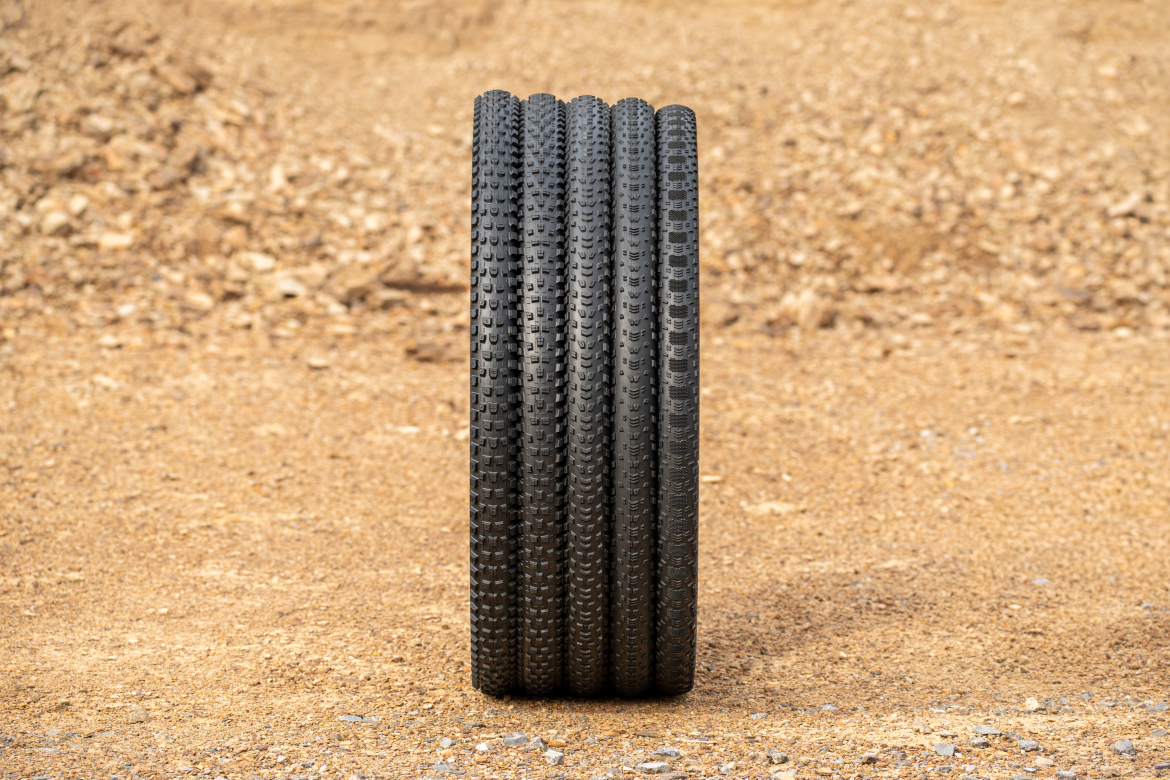 A stack of four black bicycle tires on a gravel surface, showcasing their textured tread patterns in a natural outdoor setting.