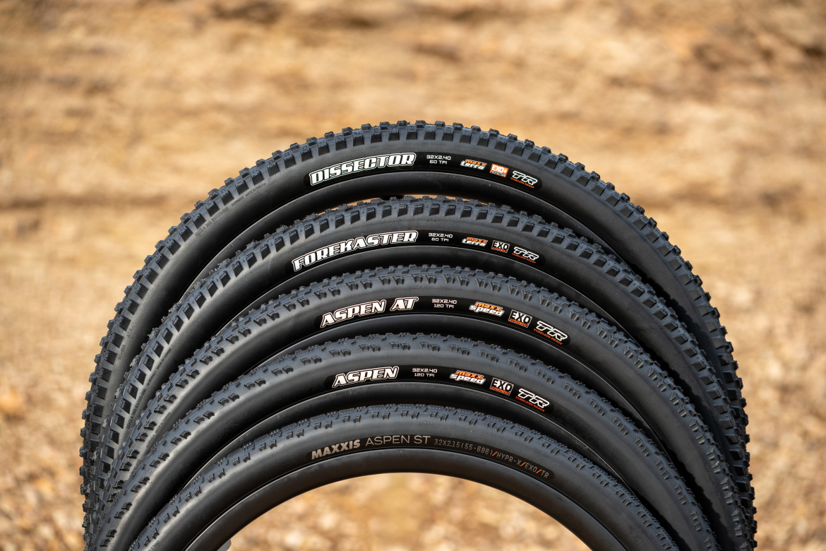 A row of five mountain bike tires arranged on a textured surface. The tires feature various tread designs and are labeled with brand names and specifications, including "Dissector," "Forekaster," "Aspen AT," "Aspen," and "Aspen ST." The background consists of a blurred, earthy texture, emphasizing the tires' details and designs.