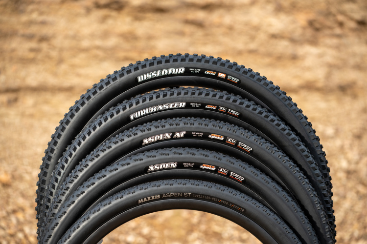 A row of five mountain bike tires arranged on a textured surface. The tires feature various tread designs and are labeled with brand names and specifications, including "Dissector," "Forekaster," "Aspen AT," "Aspen," and "Aspen ST." The background consists of a blurred, earthy texture, emphasizing the tires' details and designs.