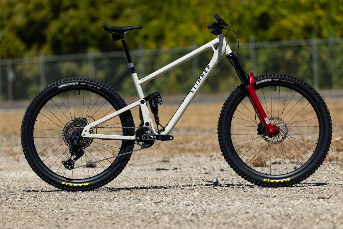 A mountain bike with a white frame and red fork is parked on a gravel surface. The bike features thick, knobby tires designed for off-road terrain, a dropper seat post, and a rear suspension system, making it suitable for trail riding. The background shows a blurred green landscape and a fence.