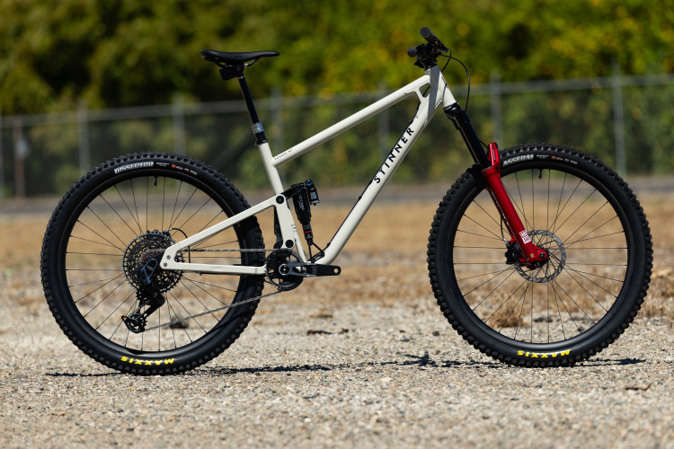 A mountain bike with a white frame and red fork is parked on a gravel surface. The bike features thick, knobby tires designed for off-road terrain, a dropper seat post, and a rear suspension system, making it suitable for trail riding. The background shows a blurred green landscape and a fence.
