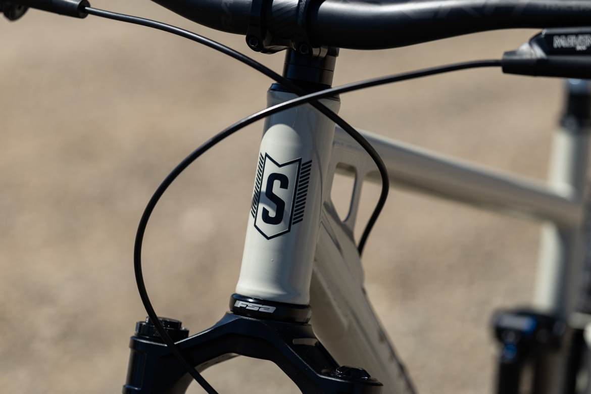 Close-up view of a bicycle's frame, showing the logo 'S' on the top tube, with black cables running from the handlebars. The components appear to be high-quality, emphasizing a sleek and modern design. The background is a blurred, light brown surface.