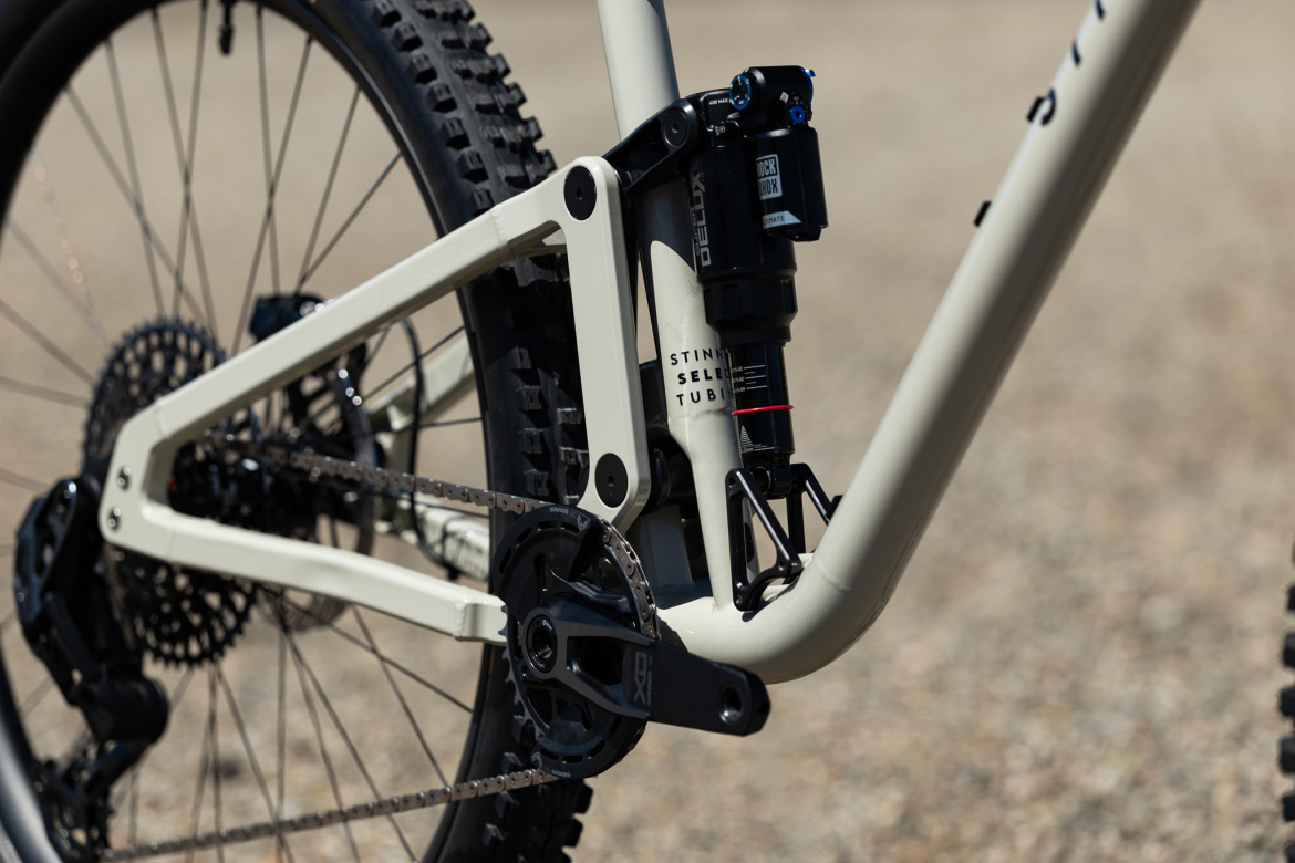 Close-up of a mountain bike's rear suspension and drivetrain. The bike features a beige frame, a visible shock absorber, and a cranks set with a chainring. The background is a rugged outdoor surface.