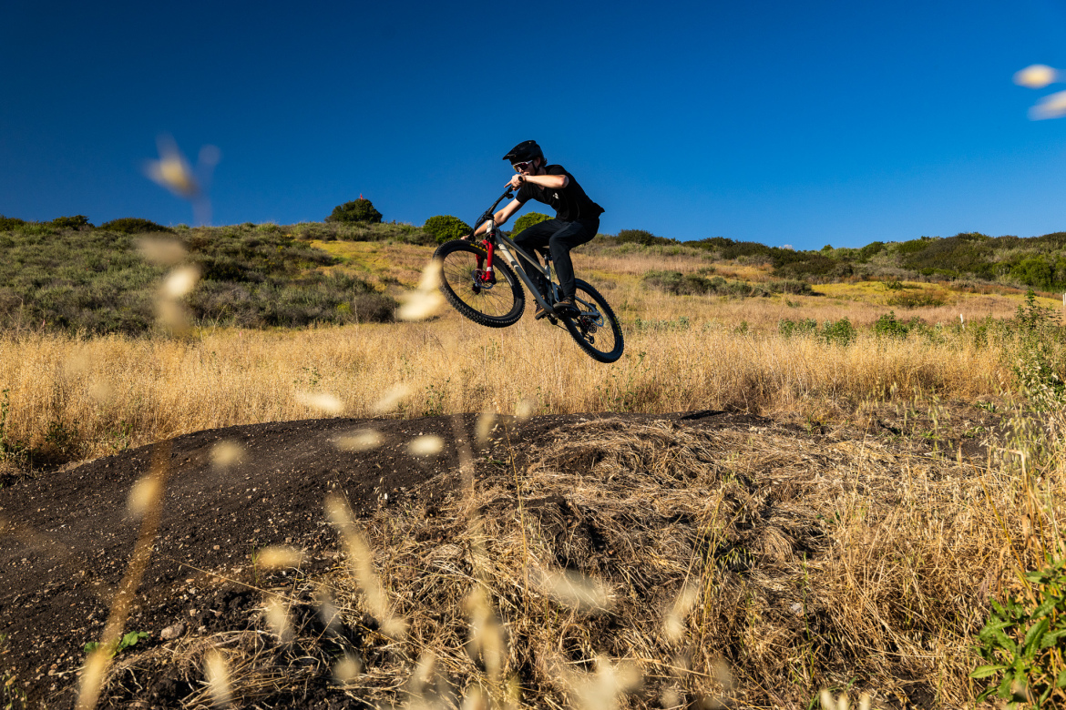 A mountain biker performs a jump over a dirt mound in a grassy outdoor setting, with a clear blue sky in the background.