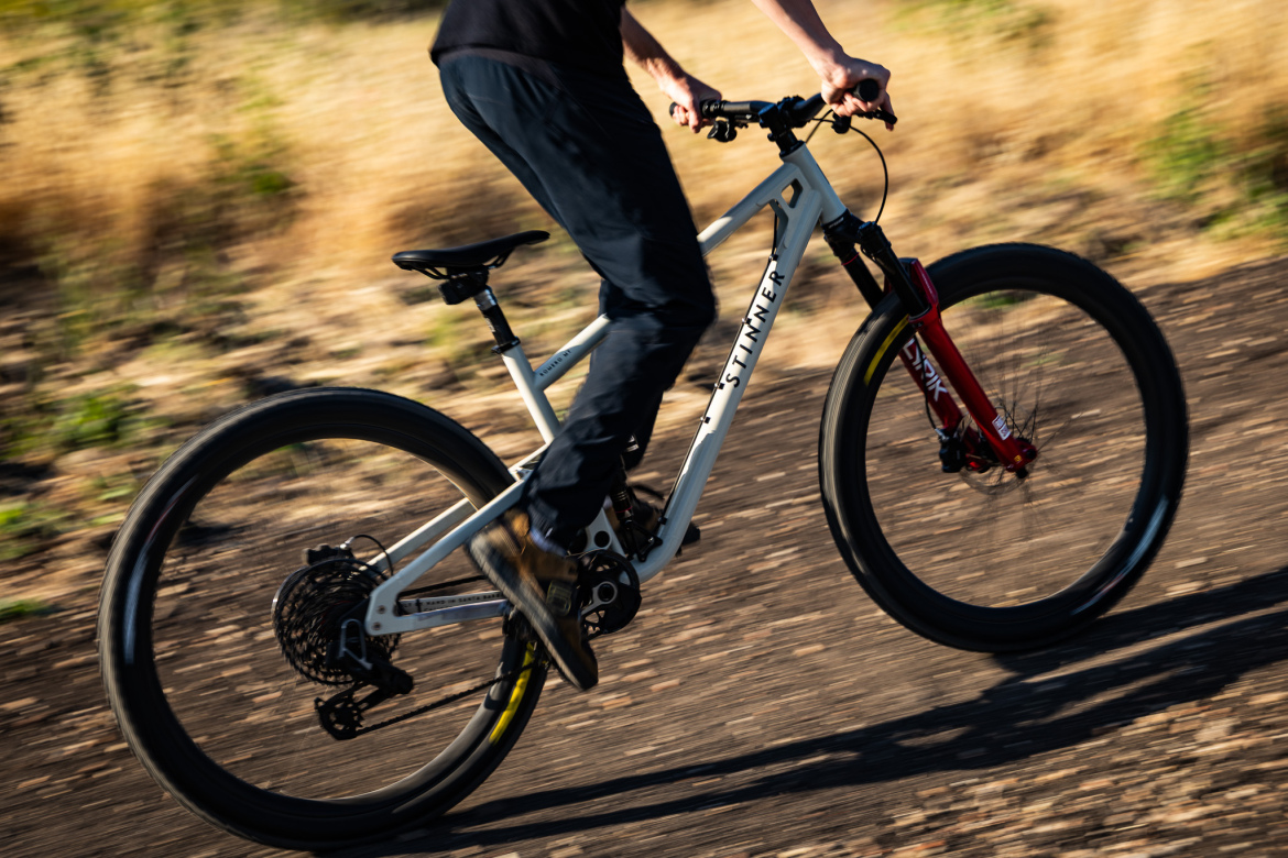 A person riding a mountain bike on a dirt trail, with the bike in motion. The bike has a light-colored frame and red front suspension forks, and the rider is wearing casual black pants and a dark shirt. The surrounding environment features dry grass and a blurred background, suggesting speed.