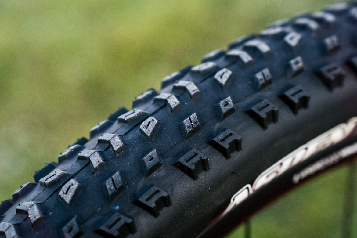 Close-up of a Maxxis Aspen AT 32" mountain bike tire showcasing its textured tread pattern, designed for traction on various terrains, with a blurred green background.