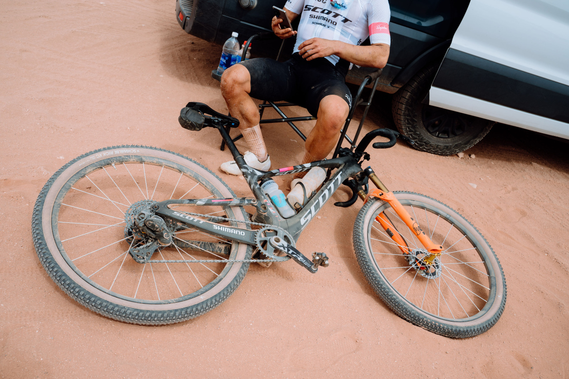 A cyclist sitting in a chair, resting with a smartphone in hand, alongside a muddy mountain bike lying on the ground. The bike has a black frame with an orange front fork and is positioned on sandy terrain, with a vehicle visible in the background.