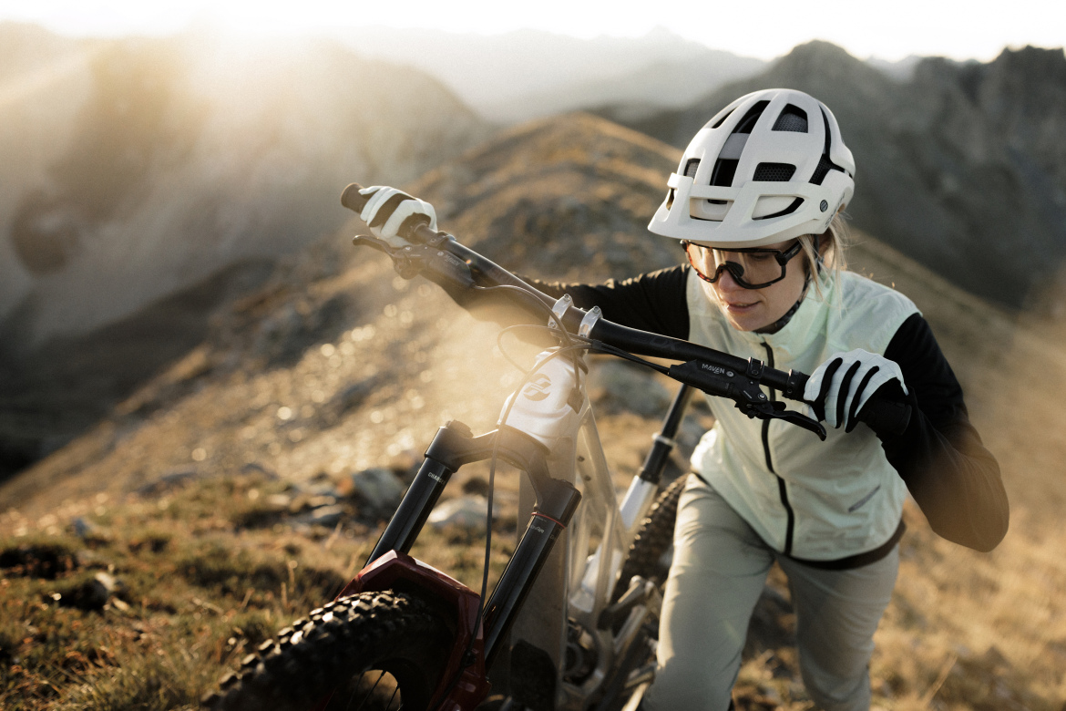 A mountain biker wearing a white helmet and sunglasses pushes their bike uphill through a rocky terrain, with sunlight illuminating the surroundings and distant mountains in the background. The rider is dressed in a light jacket and gloves, focused on navigating the challenging landscape.