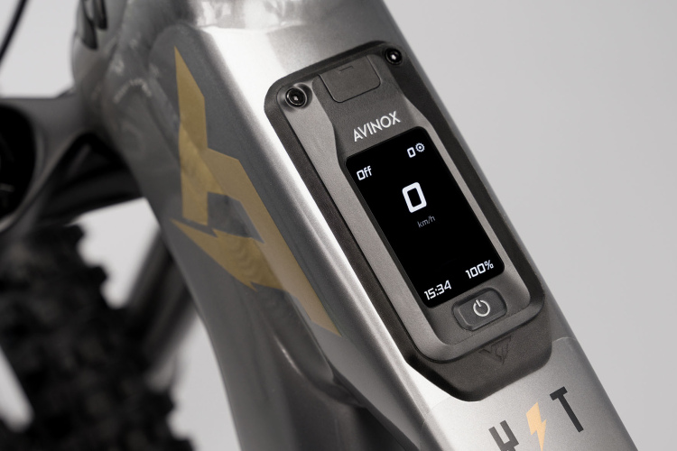 Close-up of a sleek electric bike display, showing speed as "0 km/h," battery status at "100%," and time as "15:34." The bike frame features a metallic gray finish with decorative gold accents.