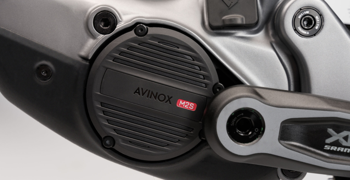 Close-up view of an AVINOX M2S motor mounted on a bicycle, featuring a sleek, metallic casing and adjacent components including a crank arm and adjustment dials.