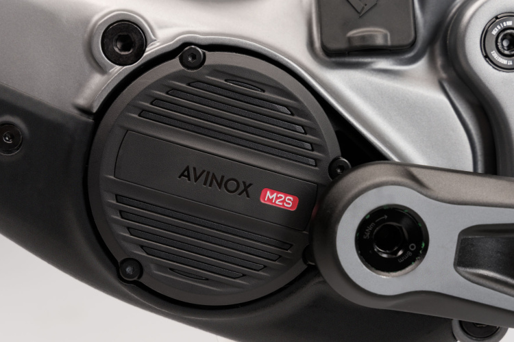 Close-up view of an AVINOX M2S motor mounted on a bicycle, featuring a sleek, metallic casing and adjacent components including a crank arm and adjustment dials.