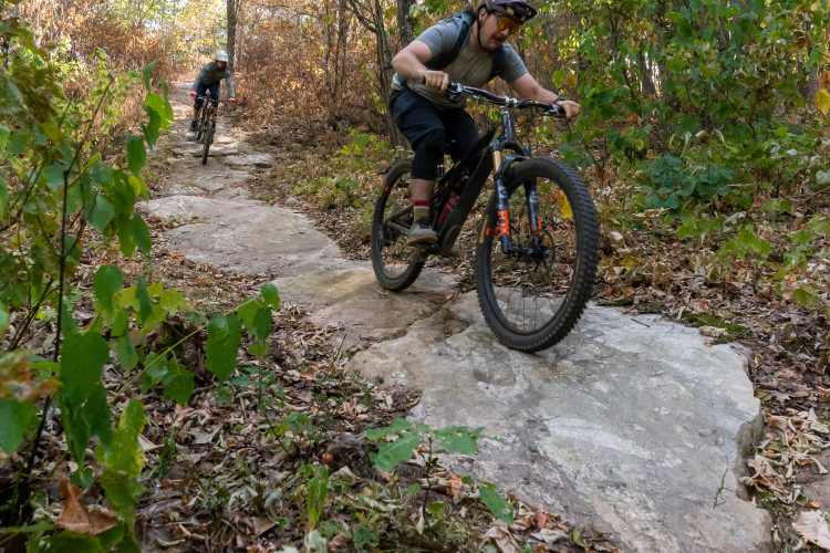 Two mountain bikers navigate a rocky trail in a wooded area during autumn. The trail is surrounded by trees with colorful foliage and scattered leaves on the ground. One rider is in the foreground, focused on navigating a boulder, while the other is further back on the trail.