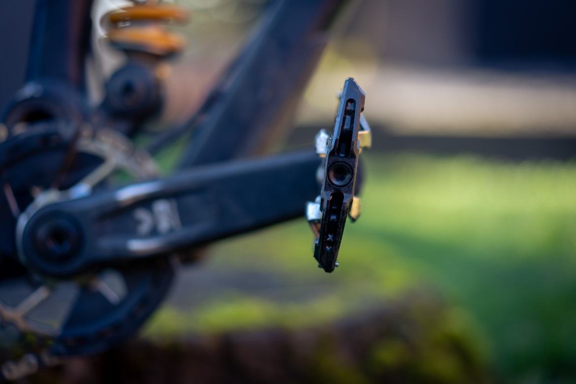 Close-up of a bicycle pedal mounted on the crank arm of a bike, set against a blurred background of green grass. The pedal features a black metallic finish with a central opening, highlighting the intricate design of the bike's drivetrain components.
