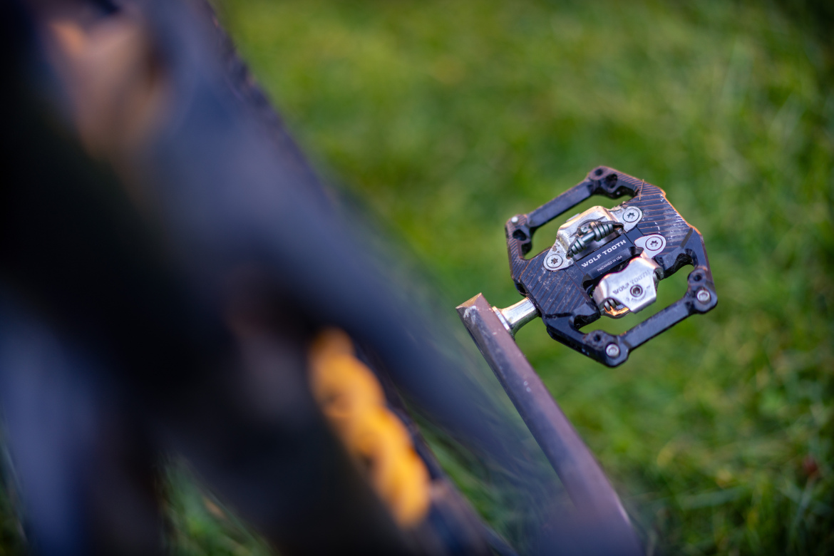 Close-up of a bike pedal featuring a textured surface, attached to a bike frame, with a blurred background of green grass. The pedal shows intricate details and is designed for securing cycling shoes.