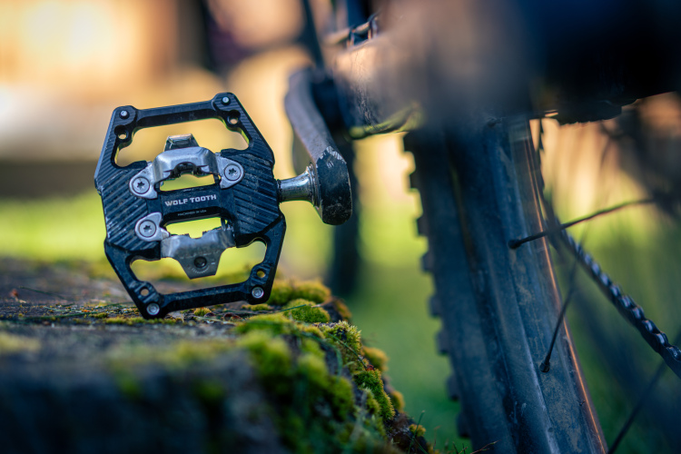 Close-up of a Wolf Tooth bicycle pedal resting on a moss-covered log, with part of a bike frame and wheel partially visible in the background, emphasizing the pedal's design and detailing.