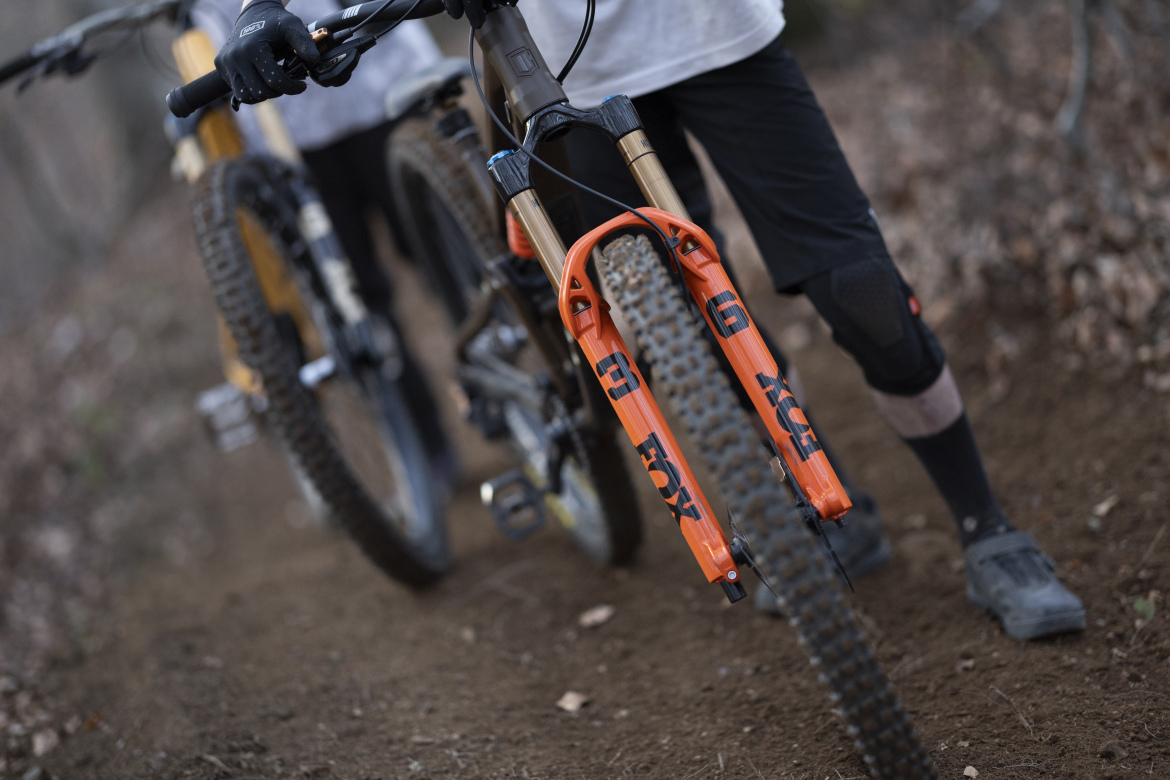 Mountain bikers walking on a dirt trail, with a close-up view of a bike's front fork featuring a vibrant orange color and the brand name "FOX." The background shows a blurred mountain bike and a wooded environment.