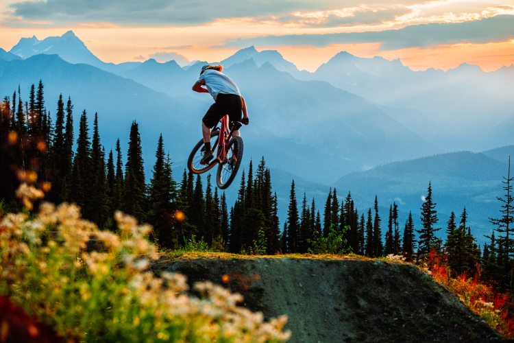 A mountain biker performing a jump over a dirt ramp, surrounded by trees and mountains at sunset. The sky is filled with soft clouds and warm colors, creating a picturesque outdoor scene.