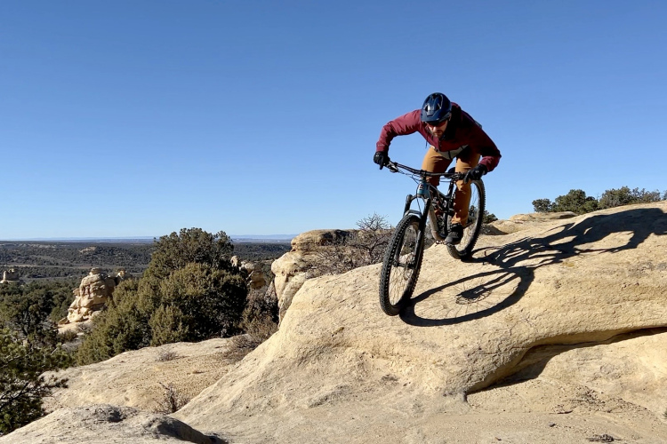 A mountain biker navigating a rocky terrain against a clear blue sky, with scenic views of the landscape in the background. The rider leans forward, skillfully maneuvering the bike over a boulder, capturing the thrill of outdoor cycling.