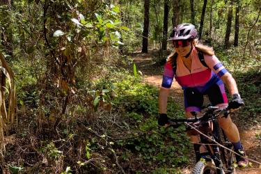 A mountain biker riding on a narrow dirt trail surrounded by lush greenery, including trees and underbrush. The cyclist is wearing a colorful cycling outfit and a helmet, focused on navigating the path. Sunlight filters through the leaves, creating dappled light on the trail. Twister mountain bike trail.