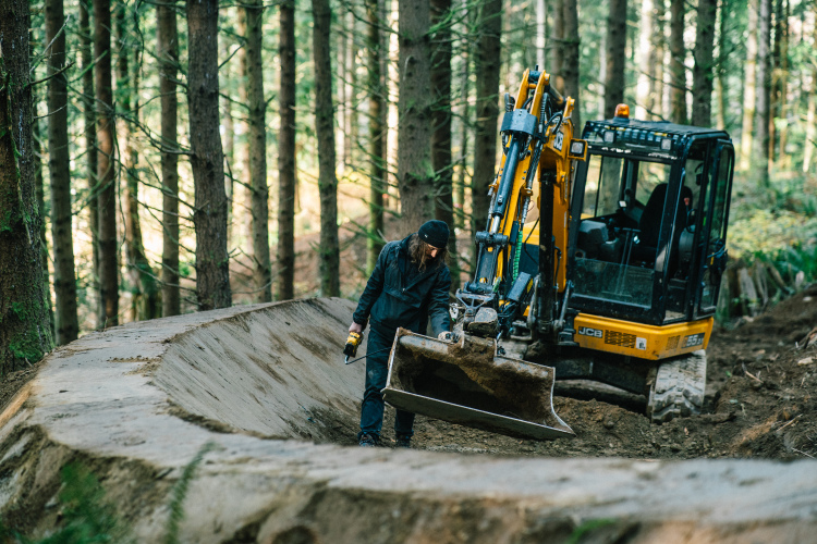 A person operating a small excavator in a forested area, working on shaping a dirt track. The individual, dressed in black clothing and wearing a beanie, is focused on their task while the excavator features a large bucket attachment. Surrounding trees and dappled sunlight create a natural backdrop.