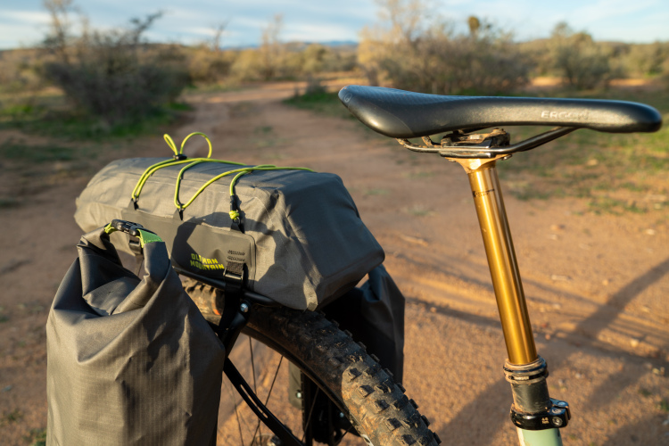 A close-up view of a bicycle with a gray cargo bag attached to the back, featuring yellow cords for securing items. The seat post is visible, showing a gold-colored dropper post and a black saddle. The bike is positioned on a dirt trail surrounded by sparse vegetation and an open landscape.