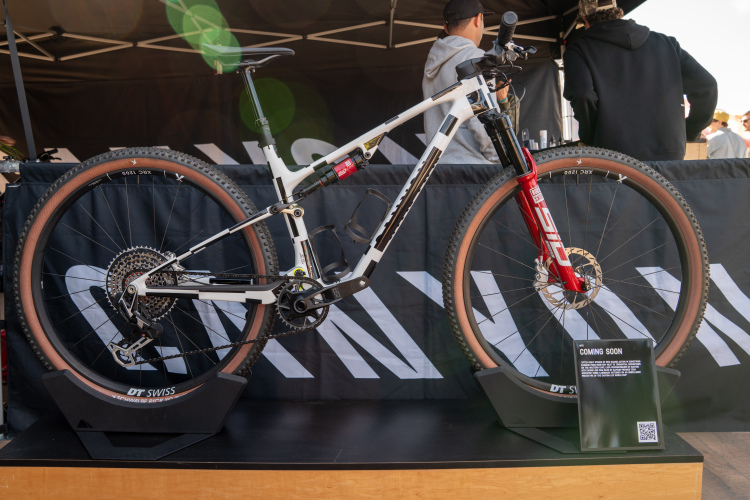 A new mountain bike displayed on a stand, featuring a white frame with black accents and a striking red front fork. The bike has brown tires and a modern design, showcasing high-performance components. In the background, a black tent with branding is visible, and a sign reads "COMING SOON," hinting at the bike's upcoming launch.