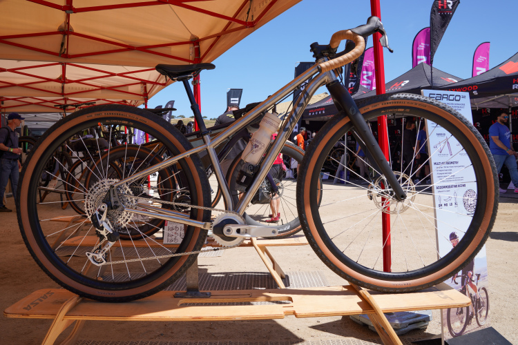 A modern bicycle displayed at an outdoor event, featuring a sleek metal frame, wide tires, and a unique design. The bike is showcased on a wooden stand under a tent, surrounded by various tents and displays in the background.