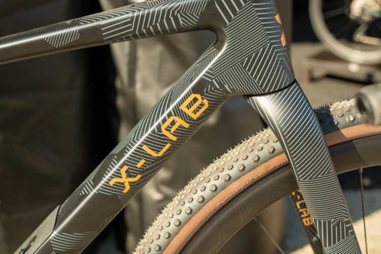 Close-up of a bicycle frame featuring a geometric design in black and gray, with the brand name "X-LAB" prominently displayed in orange. The bike has gravel tires, indicating a focus on off-road performance.