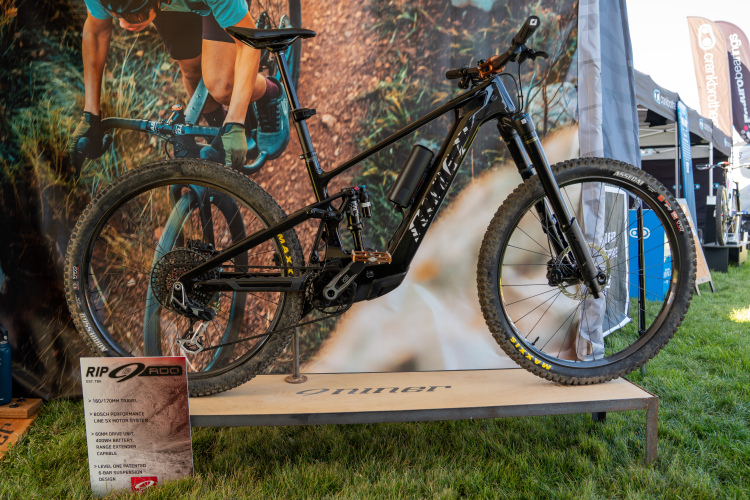 A black electric mountain bike displayed on a wooden platform, showcasing its sleek design and feature highlights. The bike features a Bosch performance motor system, 160mm/170mm travel, and a 400W battery. Behind it, there is a blurred outdoor backdrop with promotional banners visible, indicating a biking event or expo.
