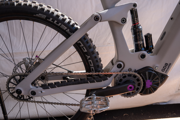 Close-up view of a mountain bike's drivetrain, showing the chain, gears, and a grey frame. The bike features a suspension shock and a visible rear tire with pronounced treads on a rugged surface. The setting includes a neutral background, suggesting it's displayed in a tent or workshop environment.