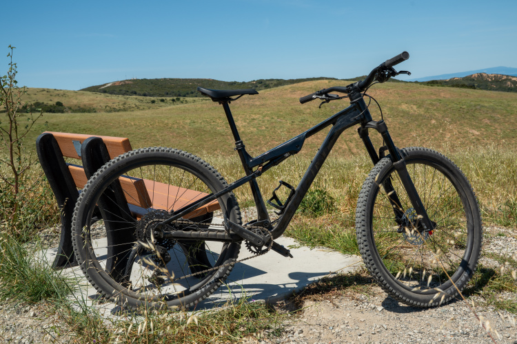 A black mountain bike is resting beside a wooden bench on a scenic hillside, surrounded by green grass and a clear blue sky. The landscape features rolling hills in the background, creating a perfect outdoor setting for cycling.