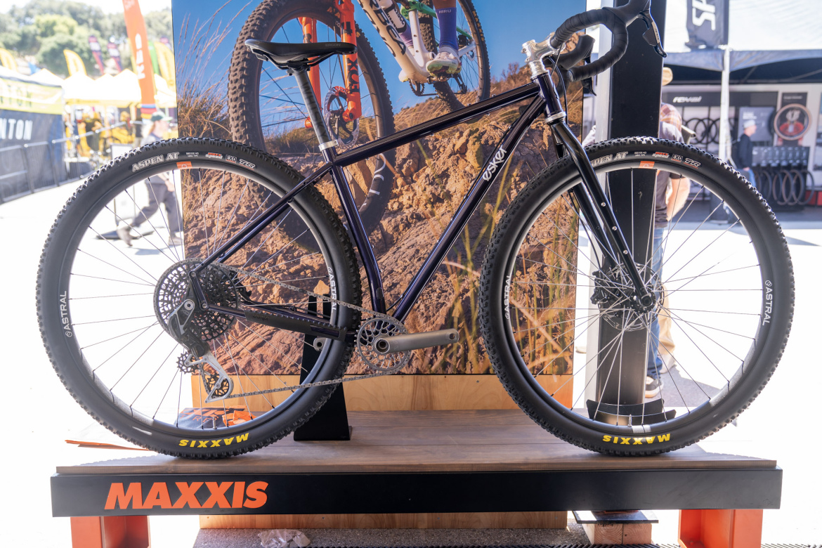 A mountain bike displayed on a stand, featuring a sleek dark frame, wide tires with Maxxis branding, and a modern gear system. In the background, there is a banner showing a trail with cyclists, and the stand is labeled with the Maxxis logo. Bright yellow tents and blurred figures of people can be seen in the outdoor event setting.