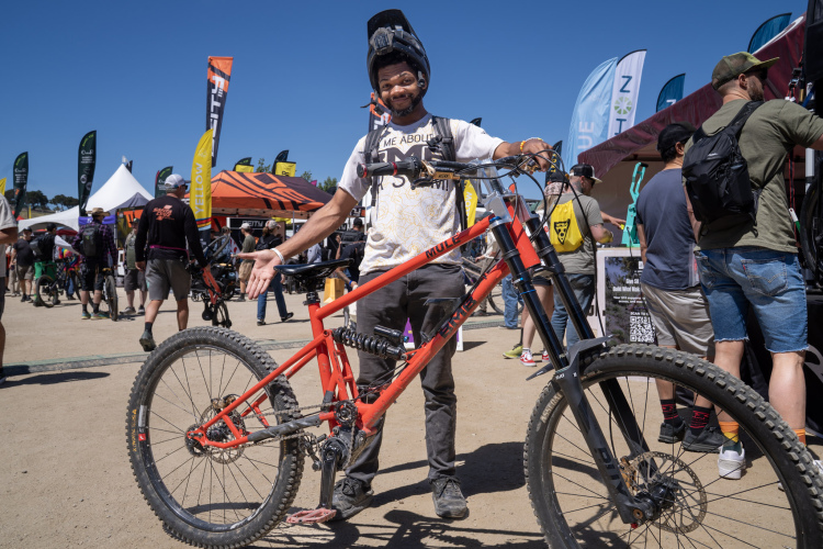 A person wearing a helmet and a casual t-shirt stands beside a bright orange mountain bike, gesturing proudly. The background shows a bustling outdoor event with various tents and banners, indicating a biking festival or expo. Other attendees can be seen exploring the area, enjoying the sunny day.