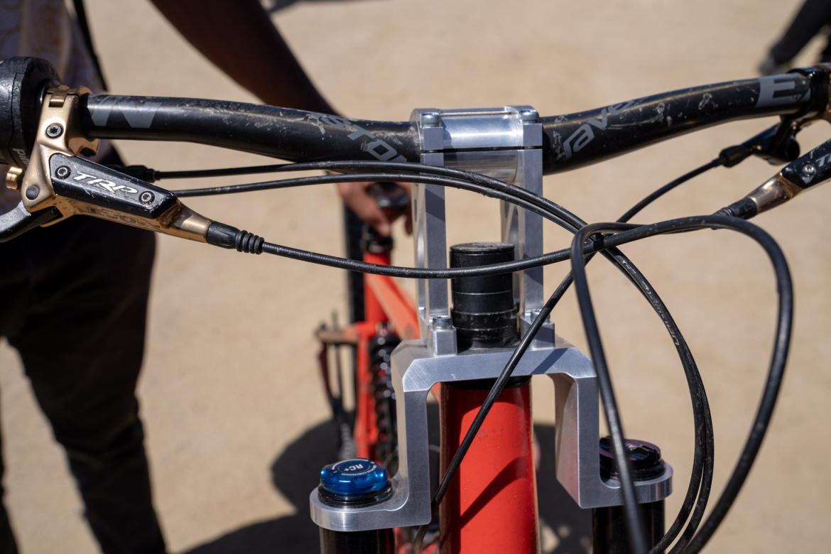 Close-up view of a mountain bike's handlebars and front fork, featuring a silver headset with cables running from the brake levers. The handlebars are black with a worn texture, and the bike frame is a bright red color, set against a sandy background.
