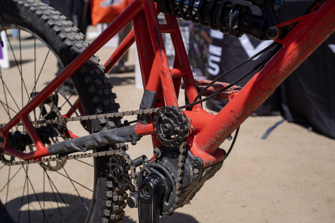 Close-up view of a red mountain bike showcasing the chain, bottom bracket, and rear suspension system. The bike appears to be on a dirt surface with some mud visible, indicating outdoor use. In the background, blurred out equipment and supplies suggest a biking event or gathering.