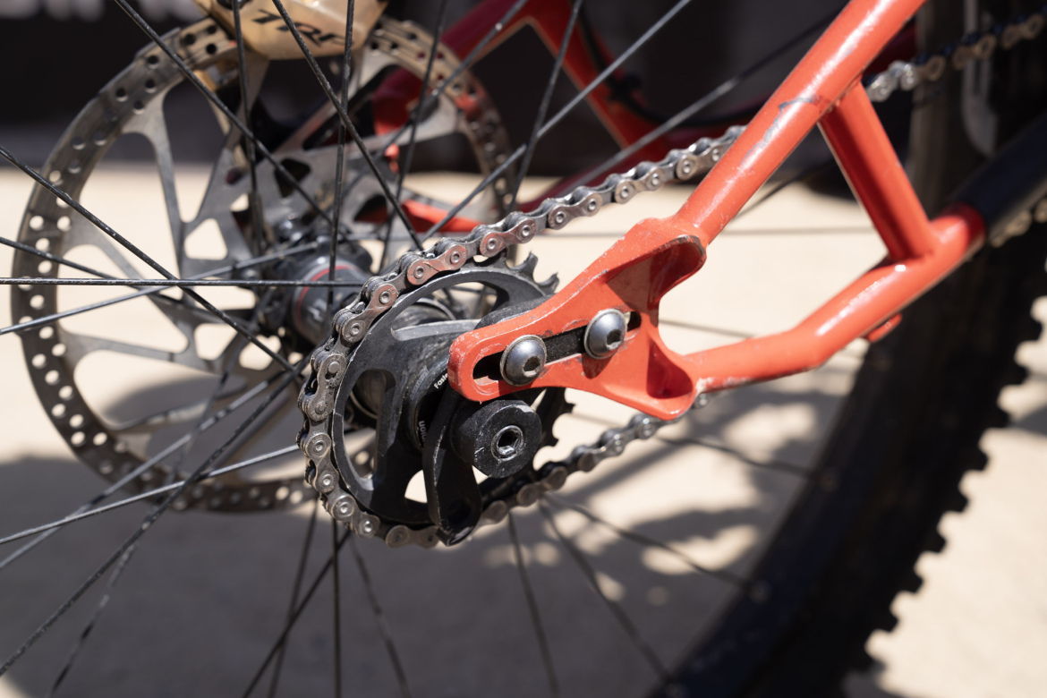 Close-up view of a bicycle drivetrain, featuring a black chain, sprocket, and a red chainstay. The image highlights the intricate components, including the disc brake and wheel spokes in the background.