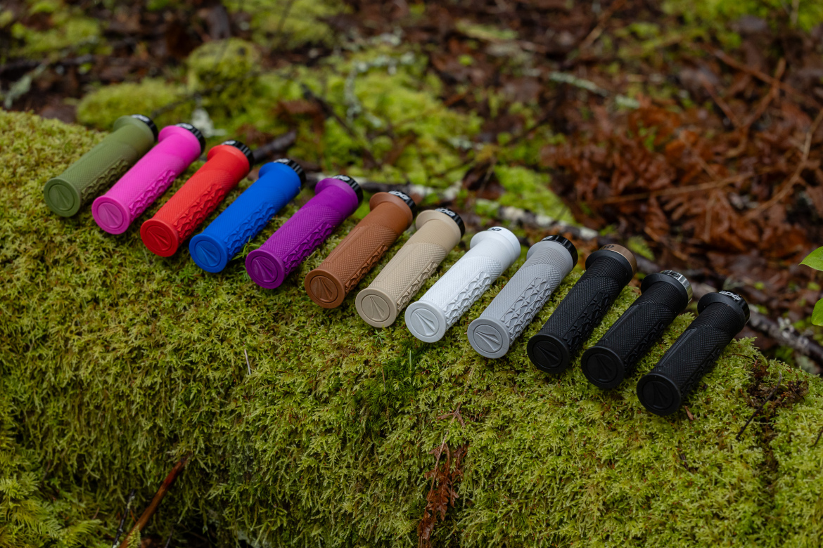 A row of twelve SDG Open Core Thrice grips displayed on a moss-covered log, showcasing various colors including green, pink, red, blue, purple, brown, tan, gray, and black. Each grip features a textured surface for improved handling. The natural forest background adds a rustic element to the image.