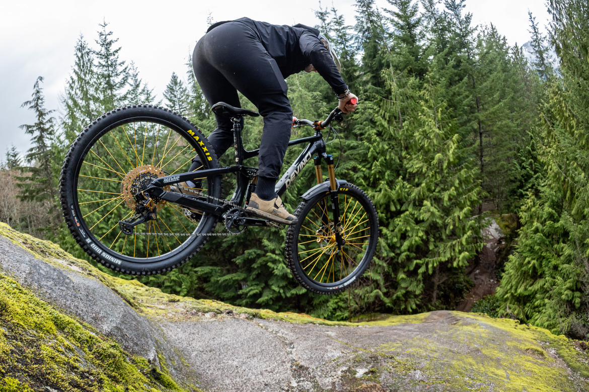 A mountain biker performing a jump off a rocky surface, surrounded by lush green trees in a nature setting. The cyclist is dressed in black riding gear and is captured mid-air, emphasizing action and adventure in mountain biking.