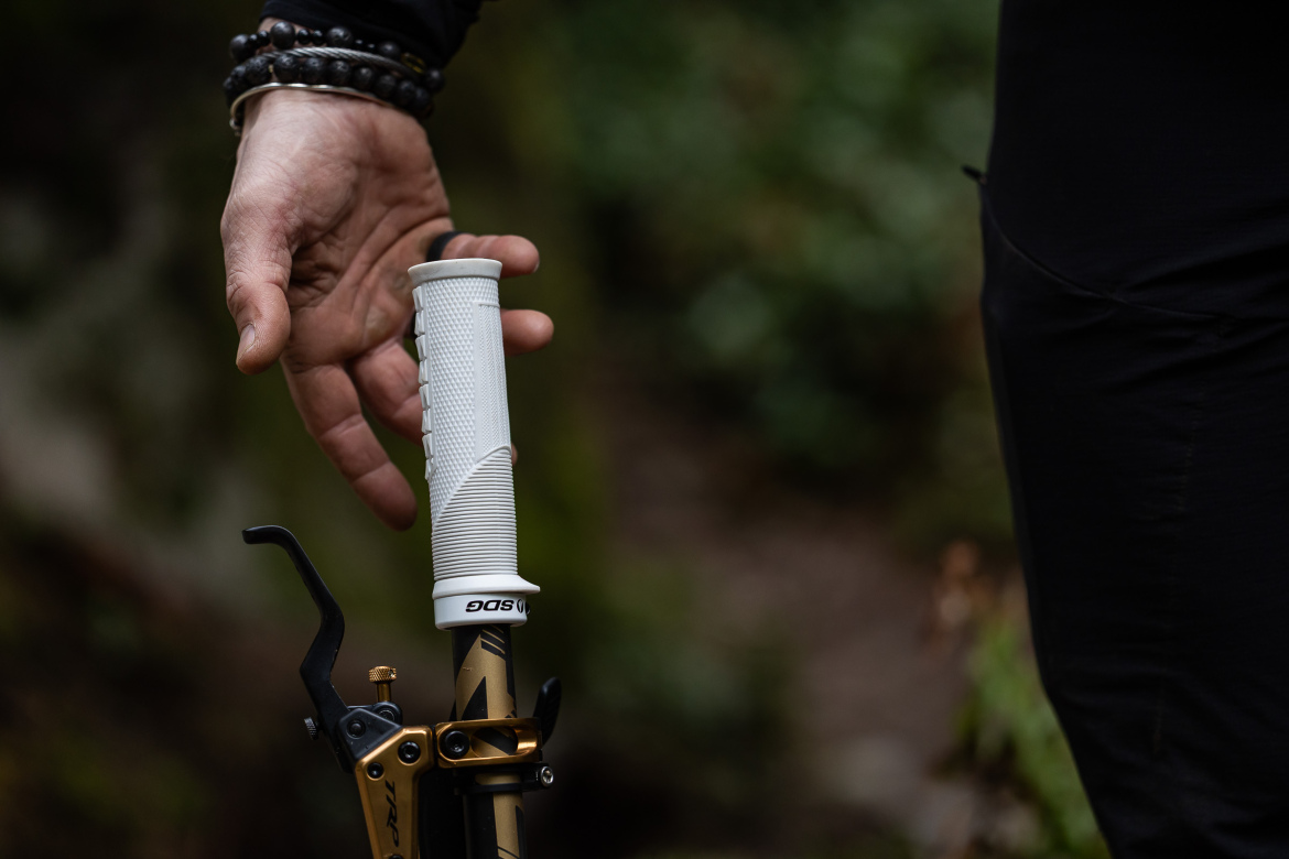 A close-up of a hand gripping the white handle of a mountain biking tool, with a blurred green background suggesting an outdoor setting. The handle features texture for improved grip, and the tool itself shows signs of gear or maintenance work. The hand is adorned with black bead bracelets, conveying an active and adventurous vibe.