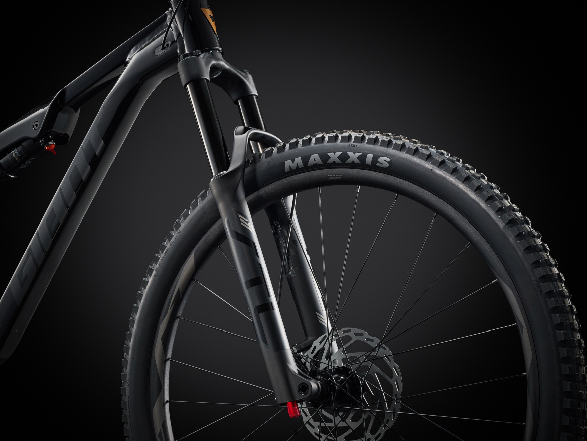 A close-up view of a mountain bike's front wheel and fork, featuring a Maxxis tire and detailed carbon frame design. The background is dark, highlighting the sleek lines and engineering of the bike components.