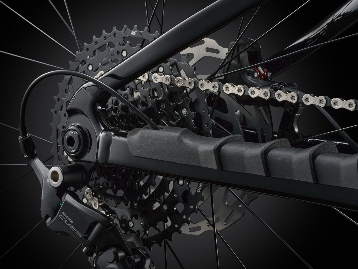 Close-up of a bicycle's rear drivetrain, showcasing the chain, cassette, and derailleur against a dark background. The image highlights the technical components, including the Shimano branding on the derailleur and the intricate details of the gear system.