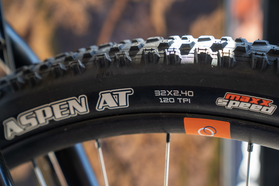 Close-up of a mountain bike tire displaying the brand name "Aspen AT" along with details including size "32X2.40" and "120 TPI." The tire is branded with "maxx speed," and the background features a blurred natural landscape.